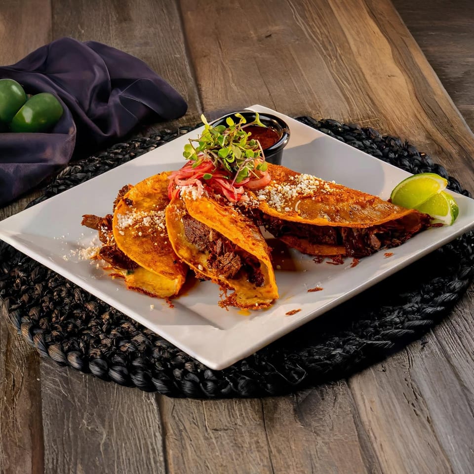 Three El Patio birria tacos with limes and sauce on a white plate, on a dark wooden table.
