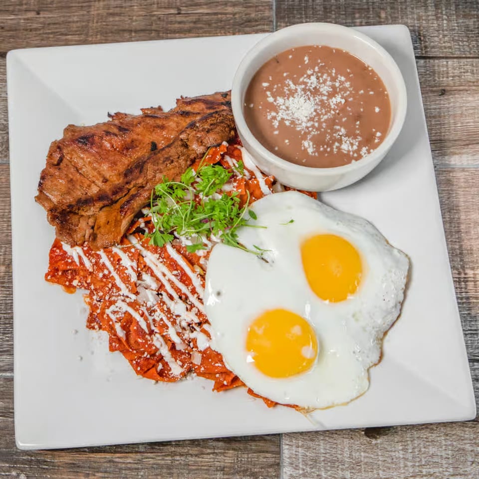 Grilled steak, chilaquiles with crema and cheese, two fried eggs, and a side of refried beans.