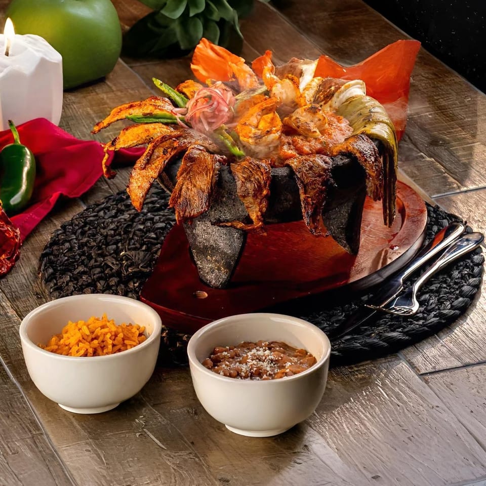 Steaming molcajete with grilled meats, shrimp, and vegetables, served with rice and beans.