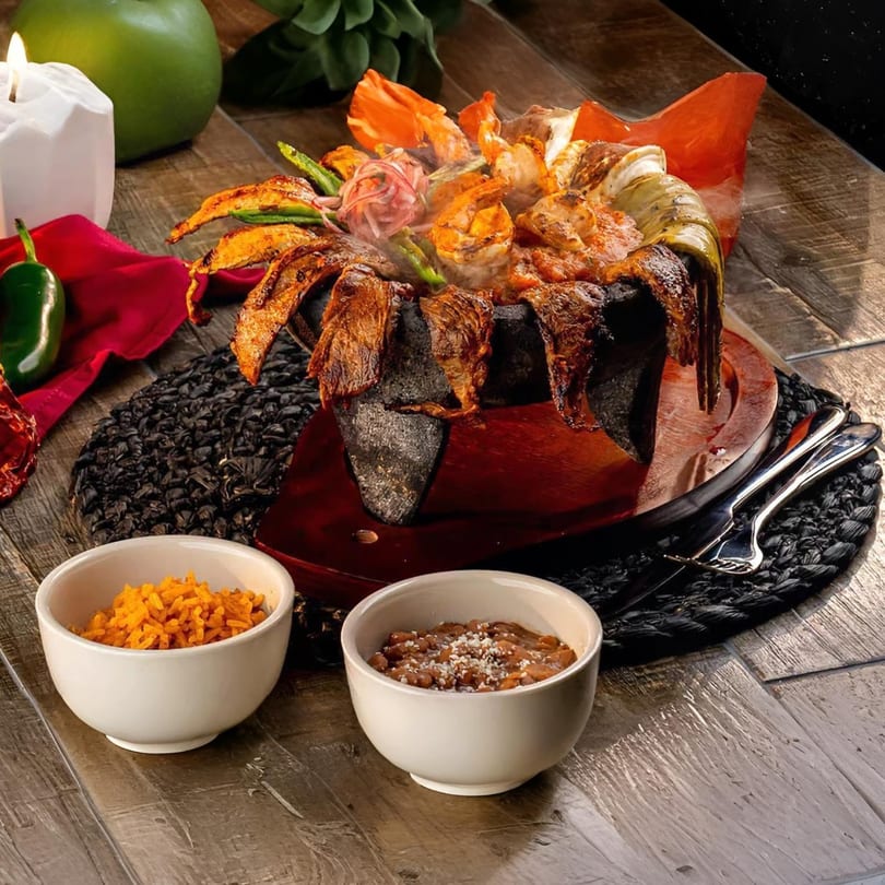 Steaming molcajete with grilled meats, shrimp, and vegetables, served with rice and beans.