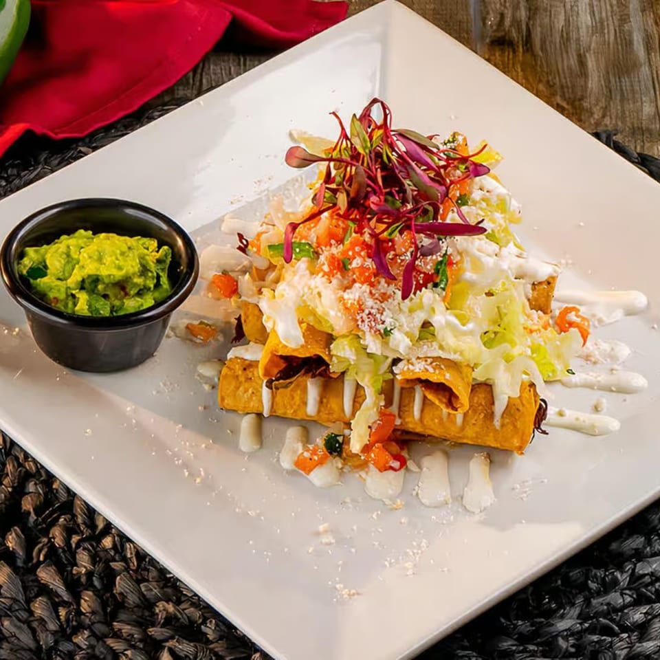 Crispy El Patio flautas topped with lettuce, pico de gallo, sour cream, and a side of guacamole.
